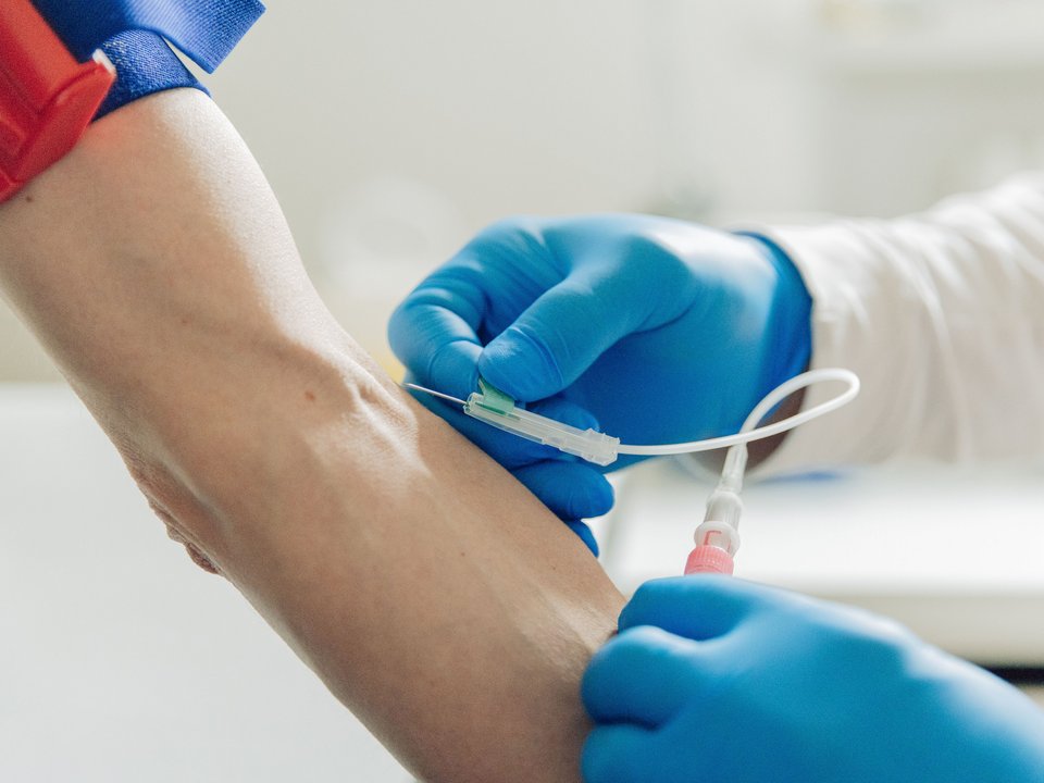 Nahaufnahme einer Blutabnahme: Fachkraft mit Schutzhandschuhen sticht Nadel in die Armbeuge.