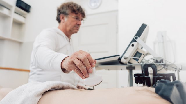 Ein Arzt führt eine Ultraschalluntersuchung durch und setzt den Schallkopf an; daneben steht das Ultraschallgerät.